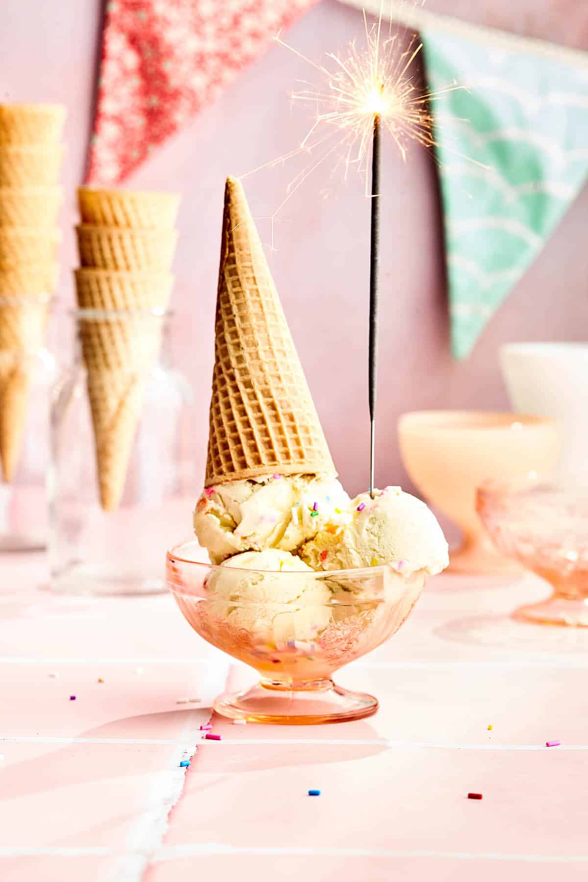Three scoops of ice cream in a pink glass dish with a sparkler and upside-down ice cream cone.