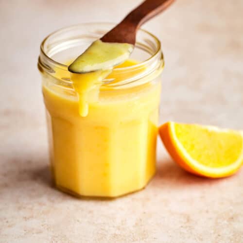 A jar of orange curd with a wooden knife on top dripping some down the side of the jar with an orange slice nearby.