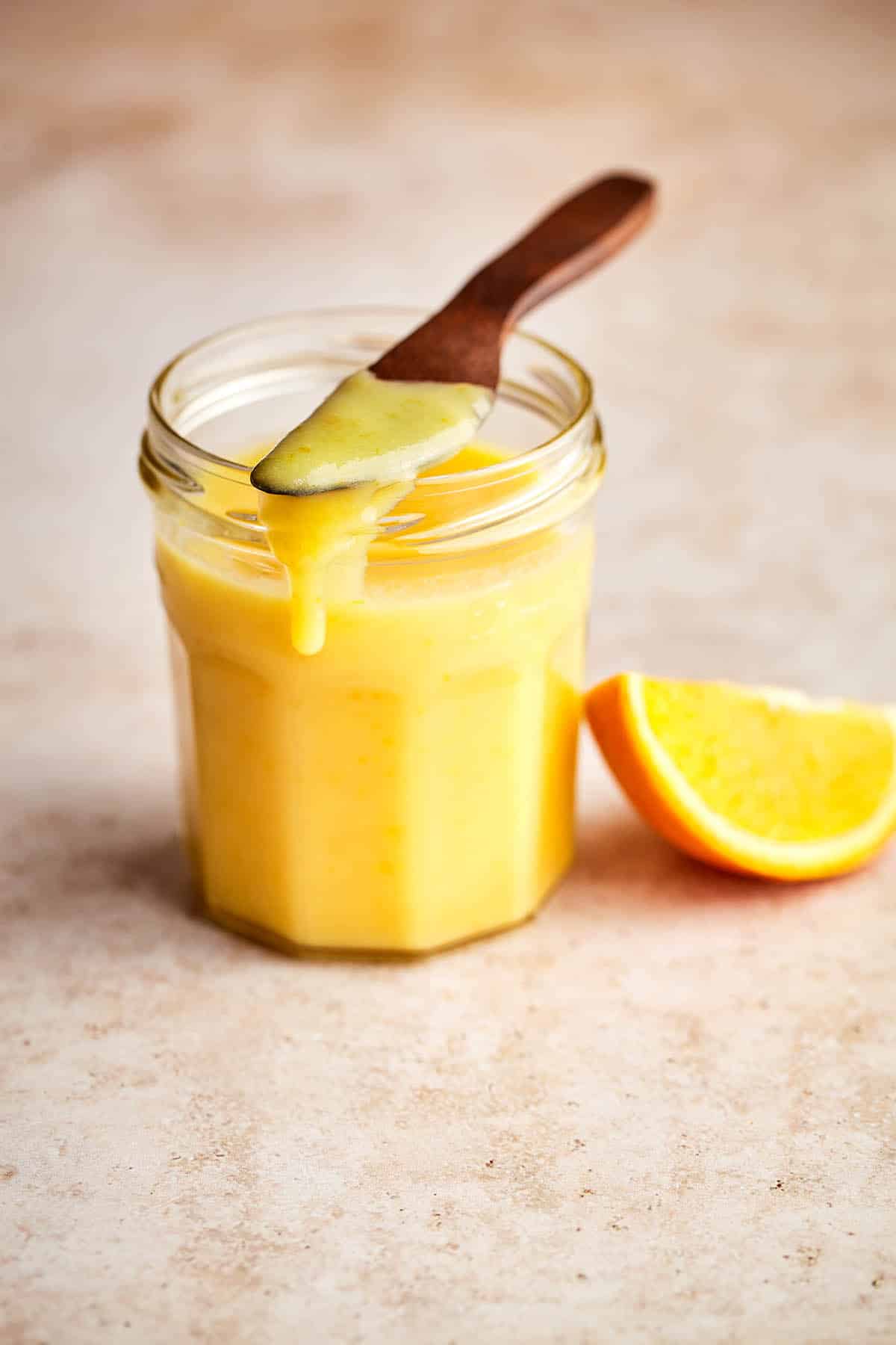 A jar of orange curd with a wooden knife on top dripping some down the side of the jar with an orange slice nearby.