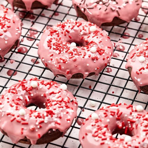 Several pink-glazed chocolate donuts with sprinkles on a wire rack.
