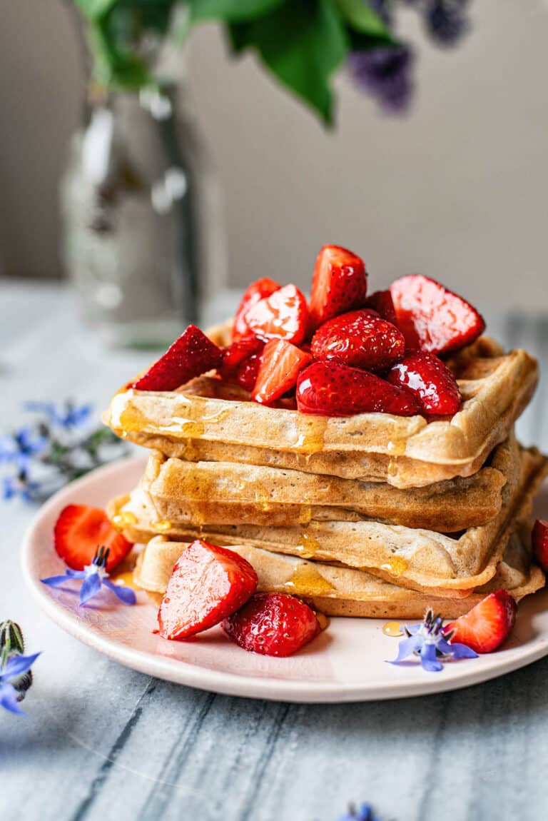 A stack of waffles on a plate, topped with strawberries and maple syrup.
