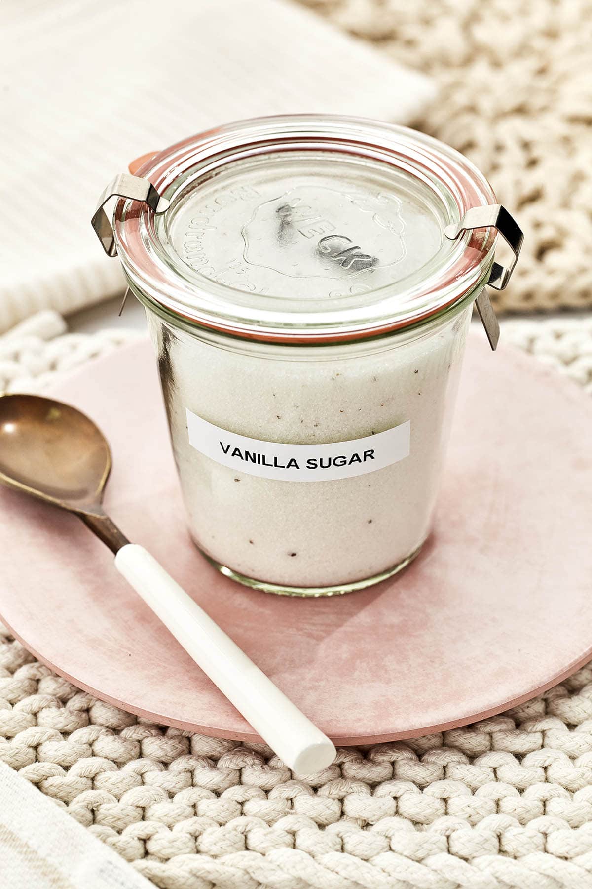 A small glass jar labeled vanilla sugar, filled with sugar, on a pink plate.