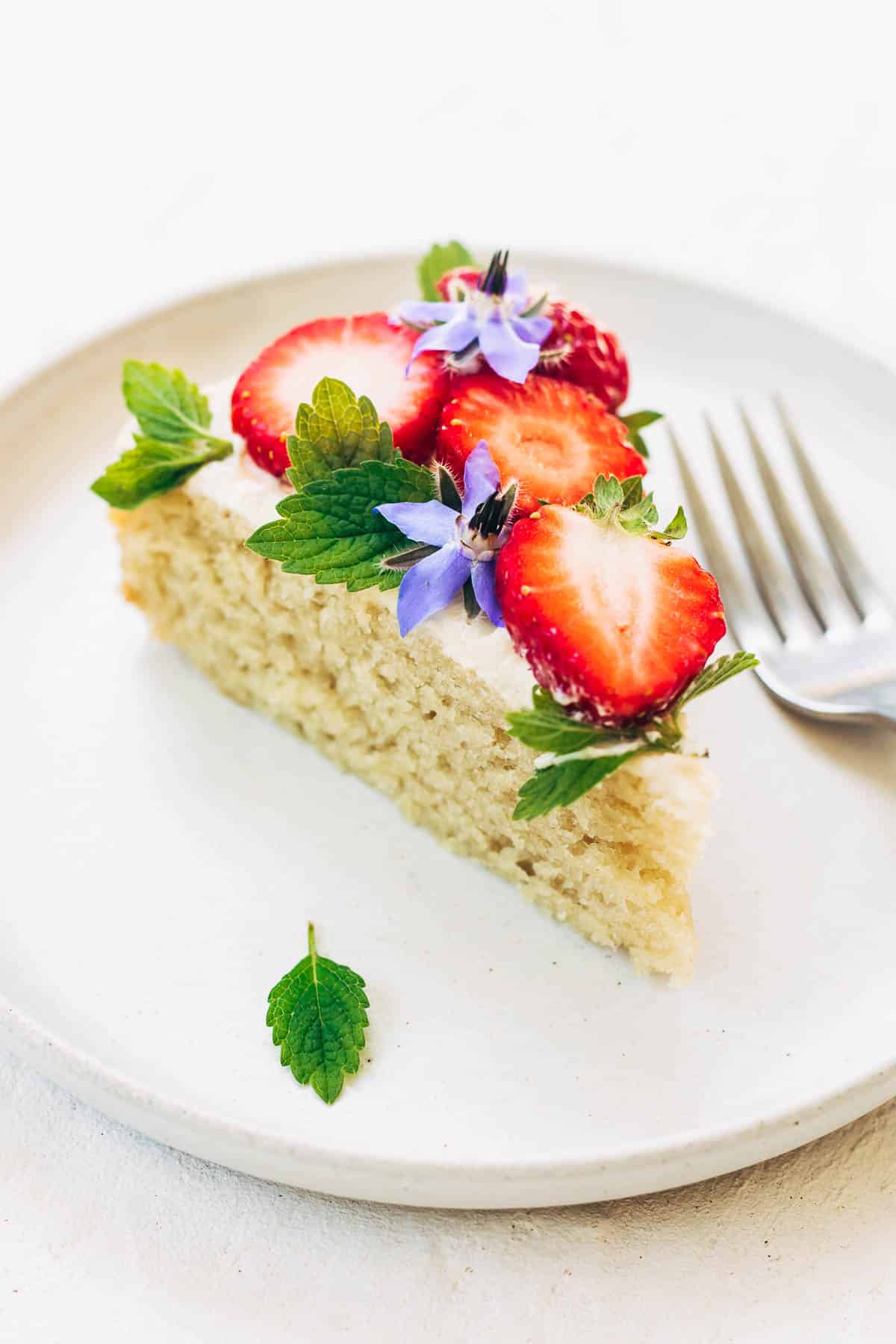 A slice of vanilla cake on a plate, topped with buttercream, fresh berries, and edible flowers.