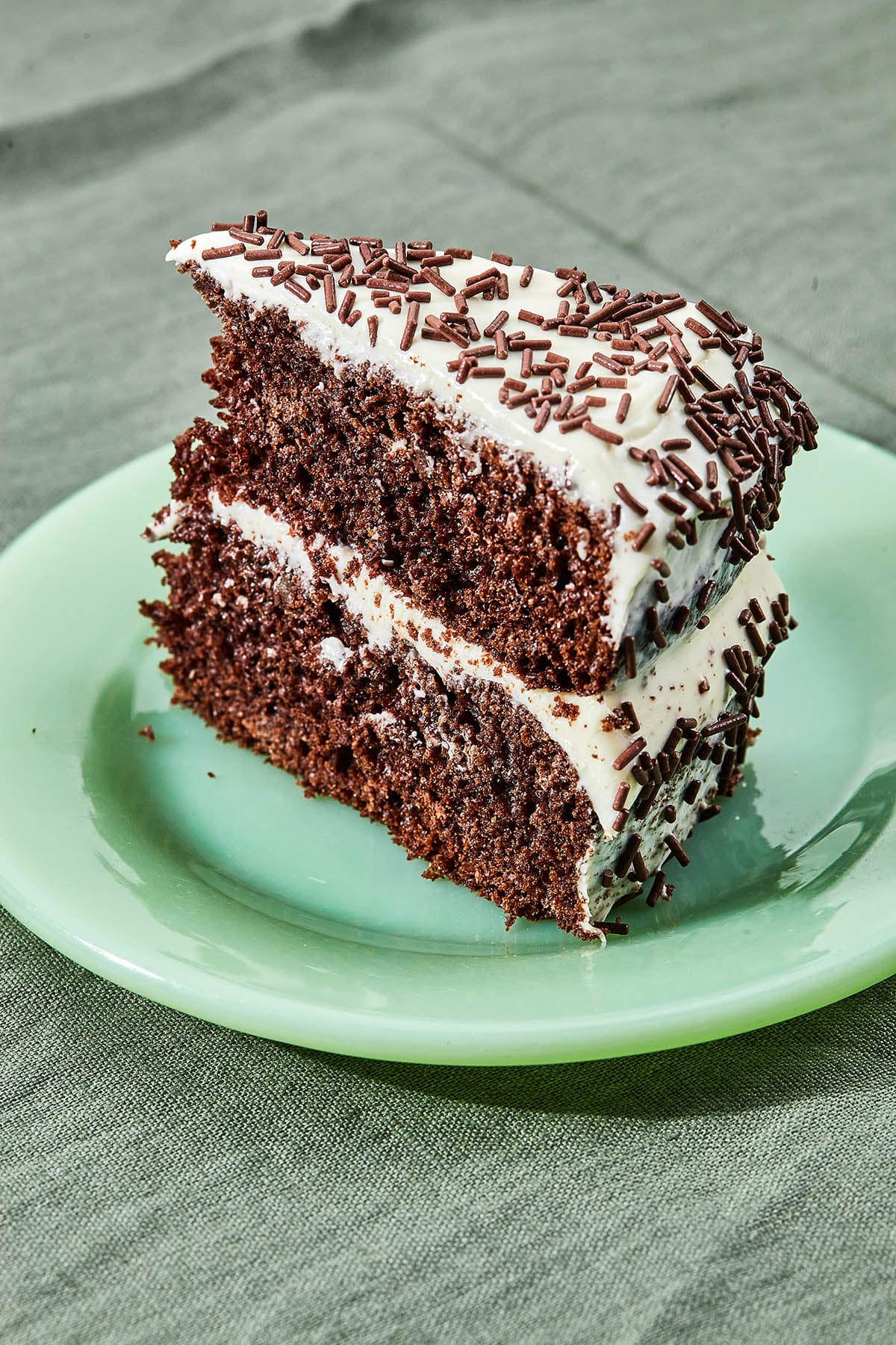 A big slice of two-layer chocolate cake with white frosting and sprinkles on a green plate.