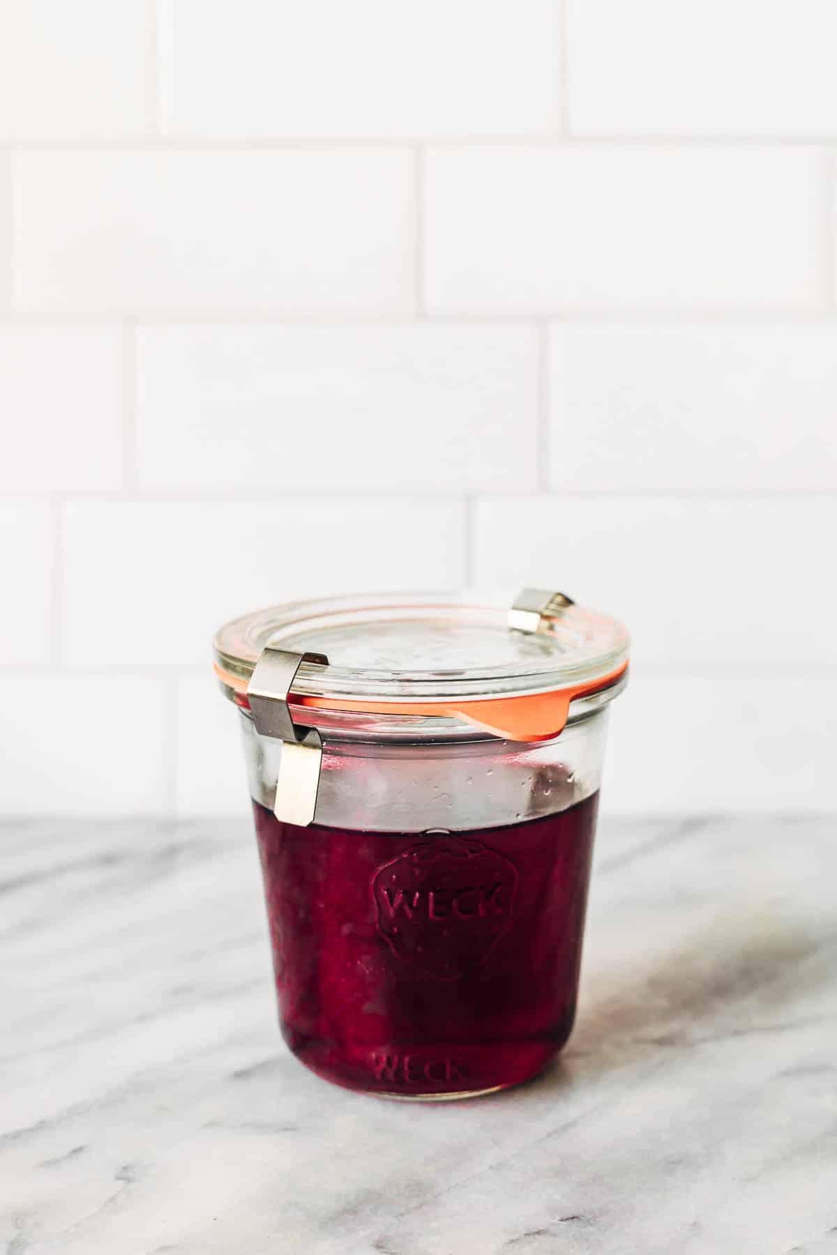 Hibiscus syrup in a small glass jar on a kitchen counter.