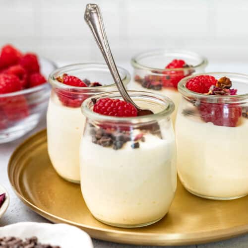 Four small glass cups of white chocolate mousse topped with raspberries.