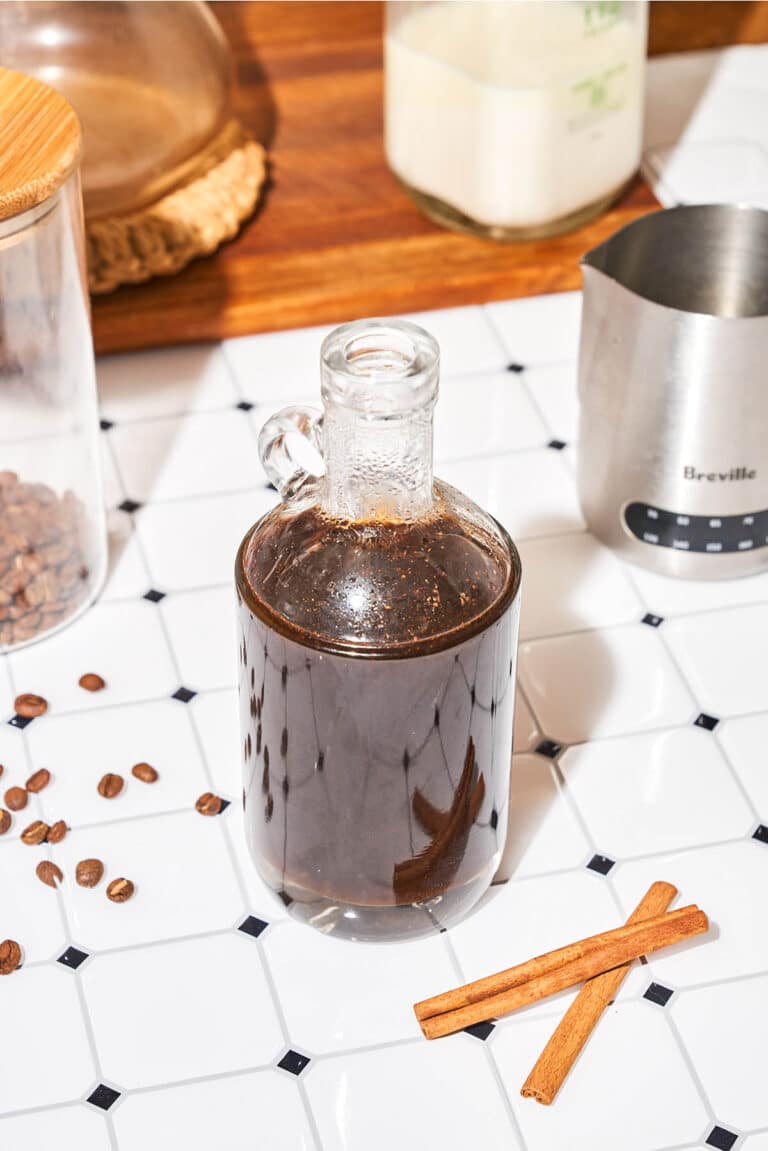 Dark brown syrup in a bottle with coffee beans and cinnamon sticks around.
