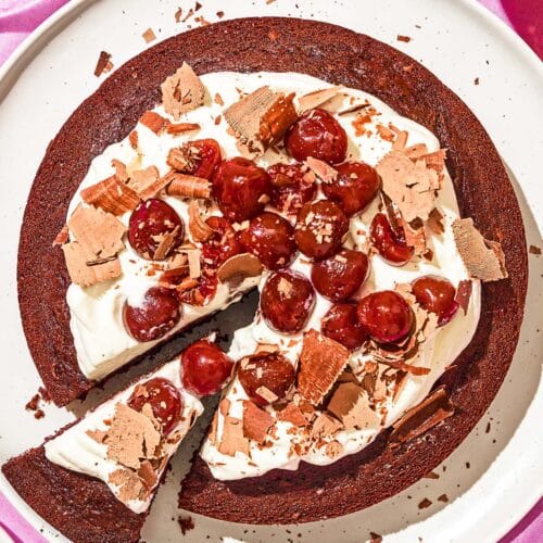 Top-down view of a chocolate cake topped with whipped cream, cherries, and chocolate shavings.