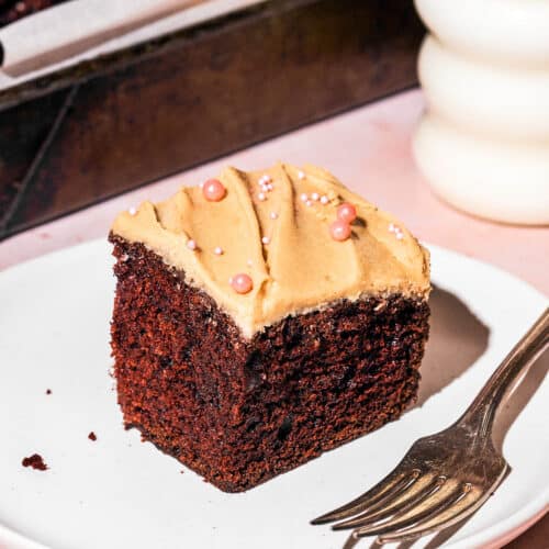 A square slice of chocolate cake with a light brown frosting on a plate.