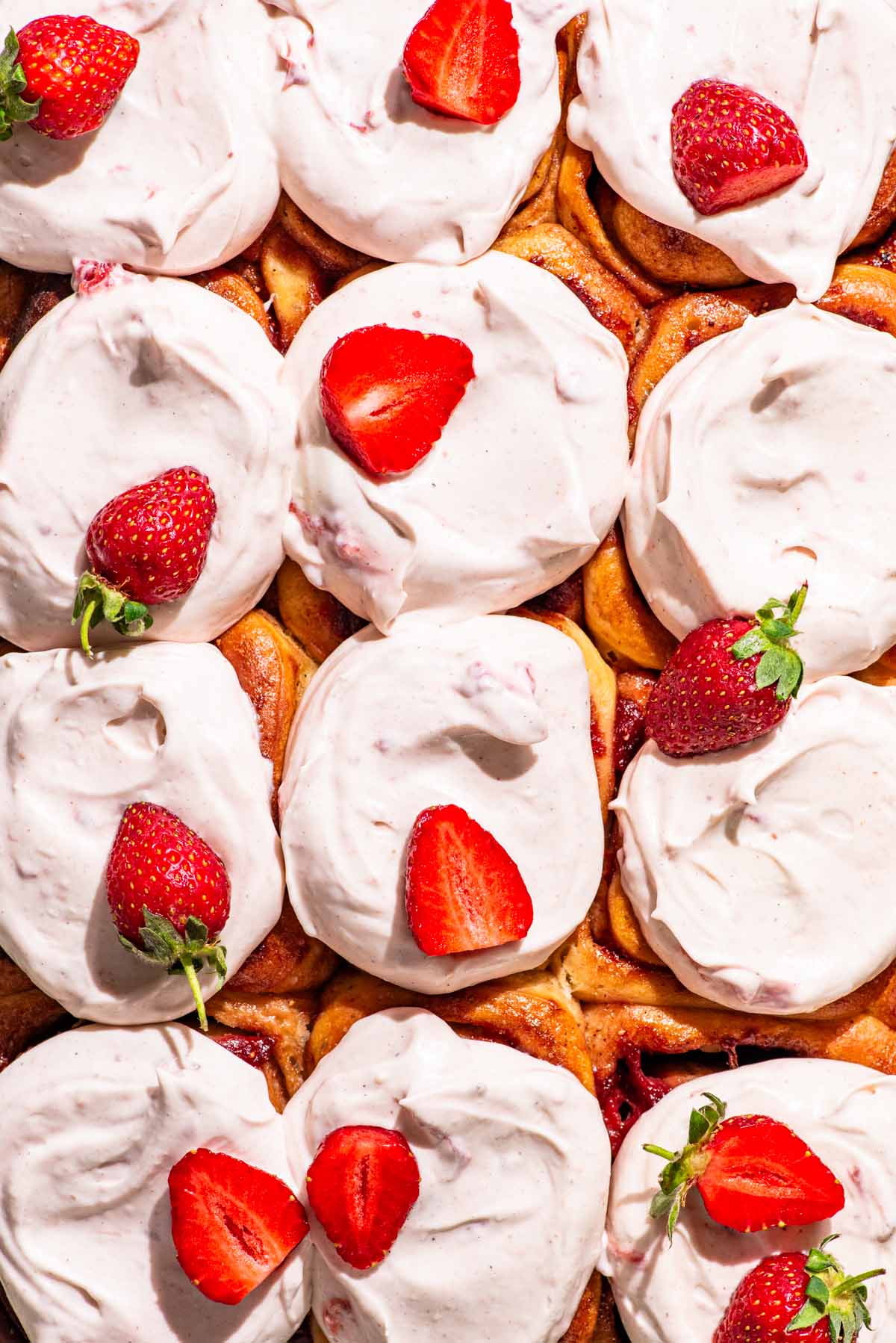 Top-down view of a batch of cinnamon rolls topped with light pink frosting and strawberries.