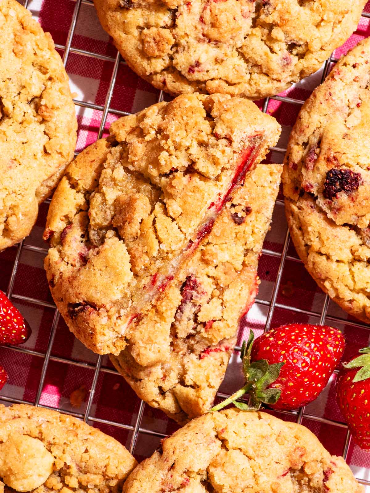 Strawberry cookies on a metal rack with fresh berries around, one cookie halved to show interior.