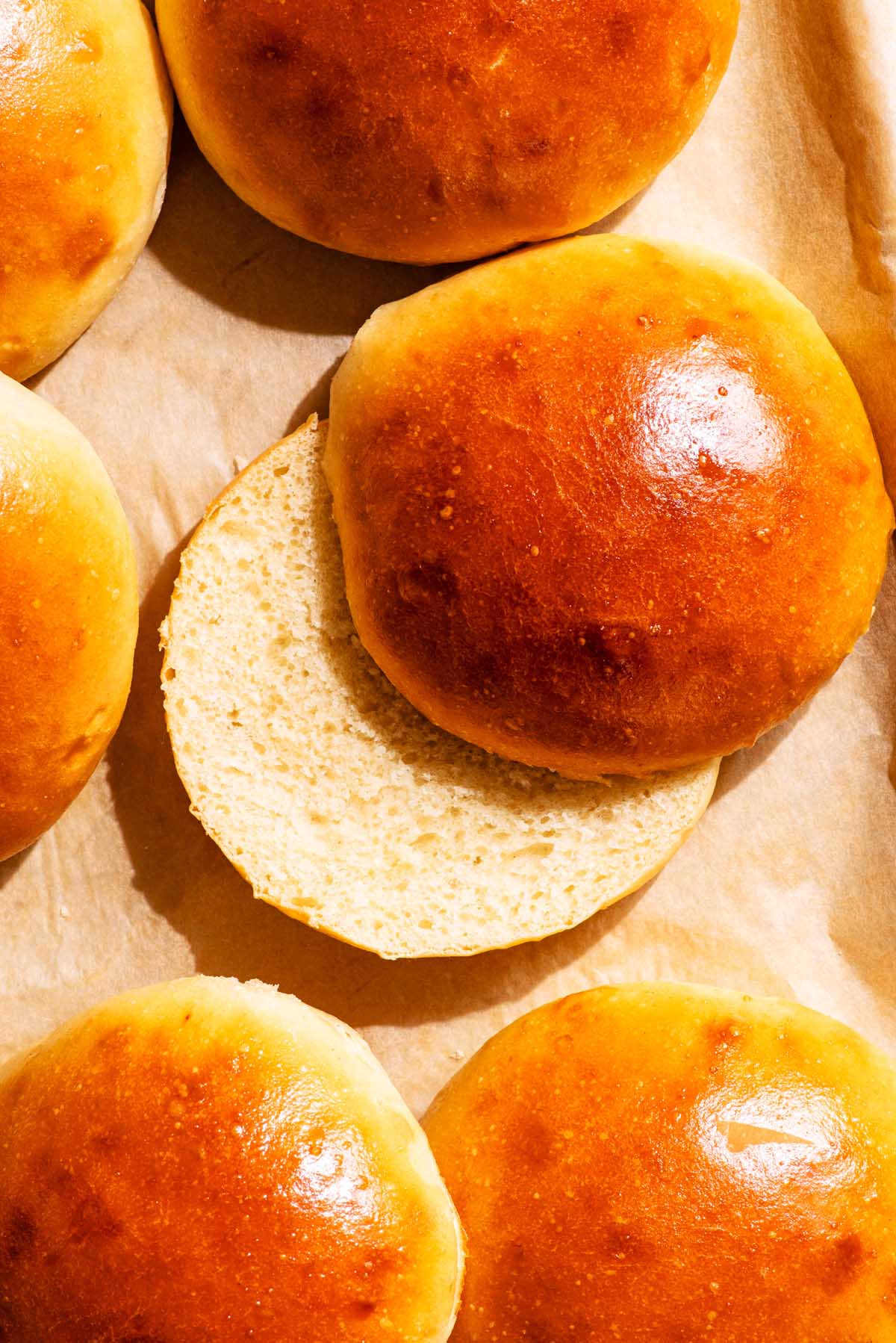 Several egg-washed buns on parchment paper, top down view, one cut to show interior texture.
