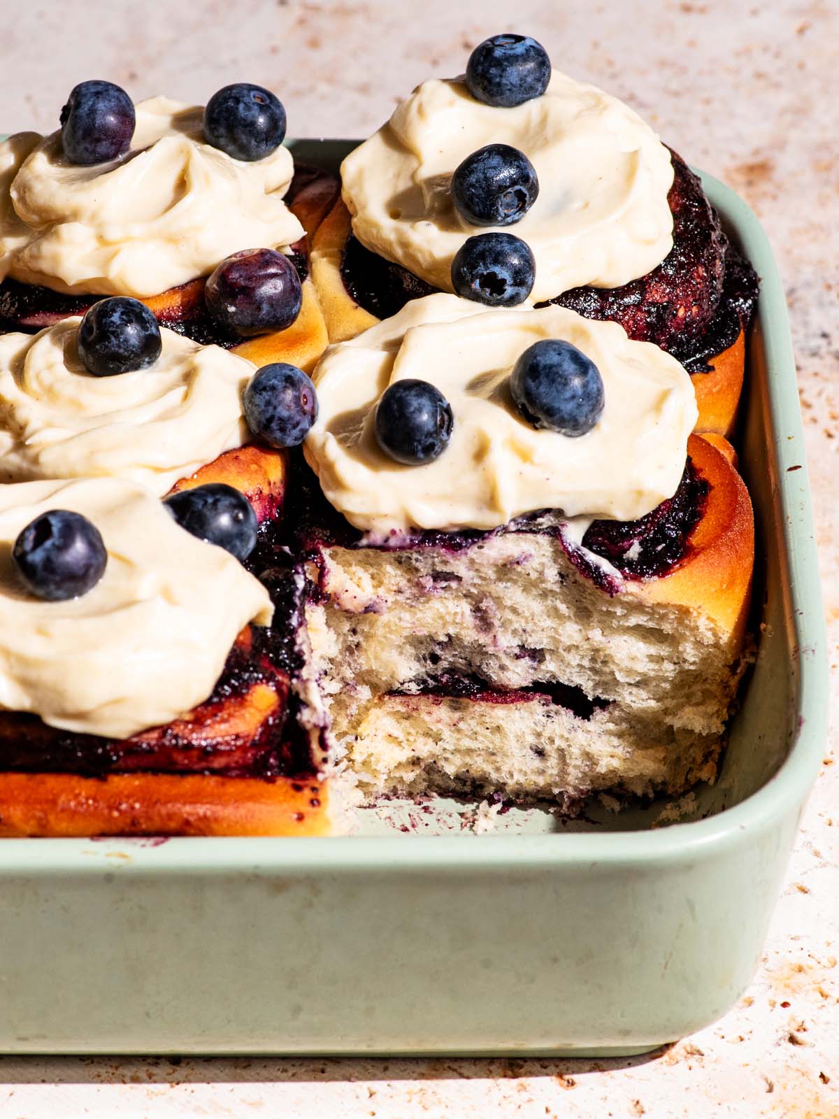 Close up of frosted cinnamon rolls with blueberries in the baking dish, one roll removed.